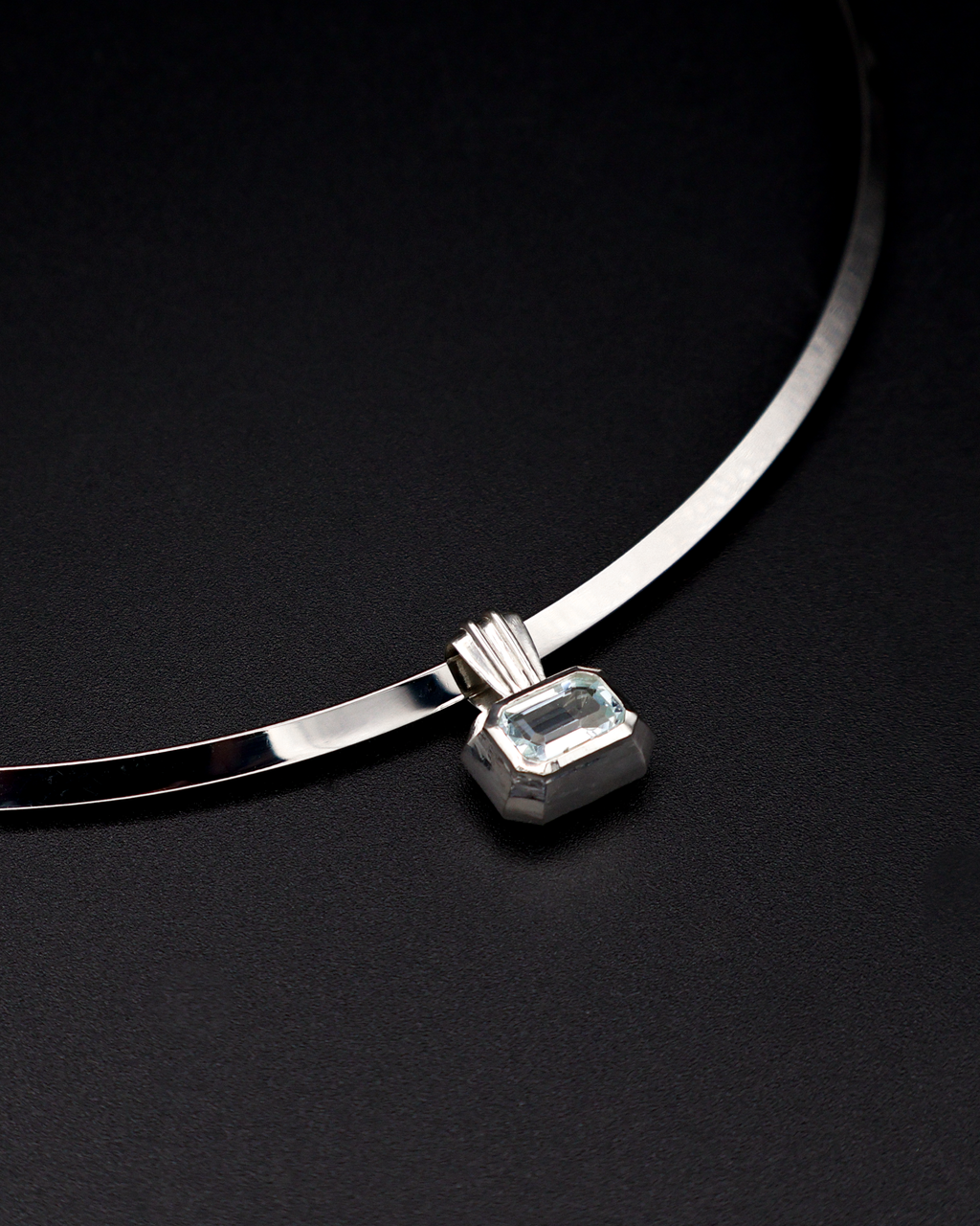 A silver-toned torque necklace with a rectangular emerald-cut aquamarine gemstone pendant, displayed on a dark textured background.