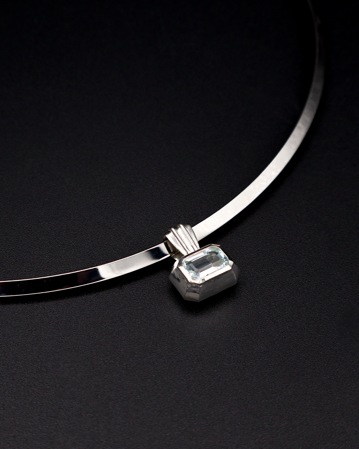 A silver-toned torque necklace with a rectangular emerald-cut aquamarine gemstone pendant, displayed on a dark textured background.