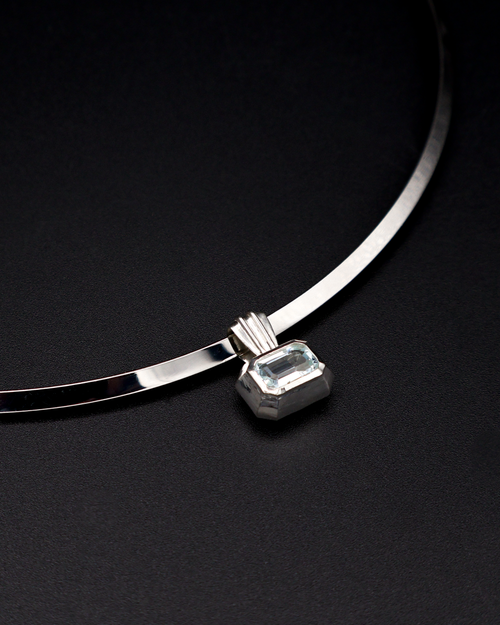 A silver-toned torque necklace with a rectangular emerald-cut aquamarine gemstone pendant, displayed on a dark textured background.